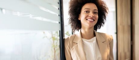 Eine lächelnde Frau in Business-Kleidung steht in einem modernen Büro und blickt freundlich in die Kamera.