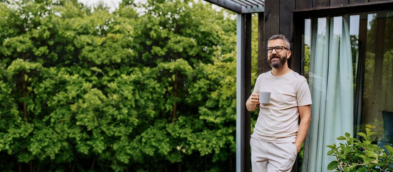 Mann steht lächelnd mit Kaffeetasse in der Hand auf der Terrasse seines Hauses im Grünen